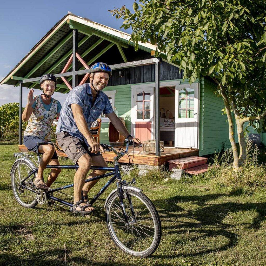 To mennesker på tandemcykel foran lille hytte